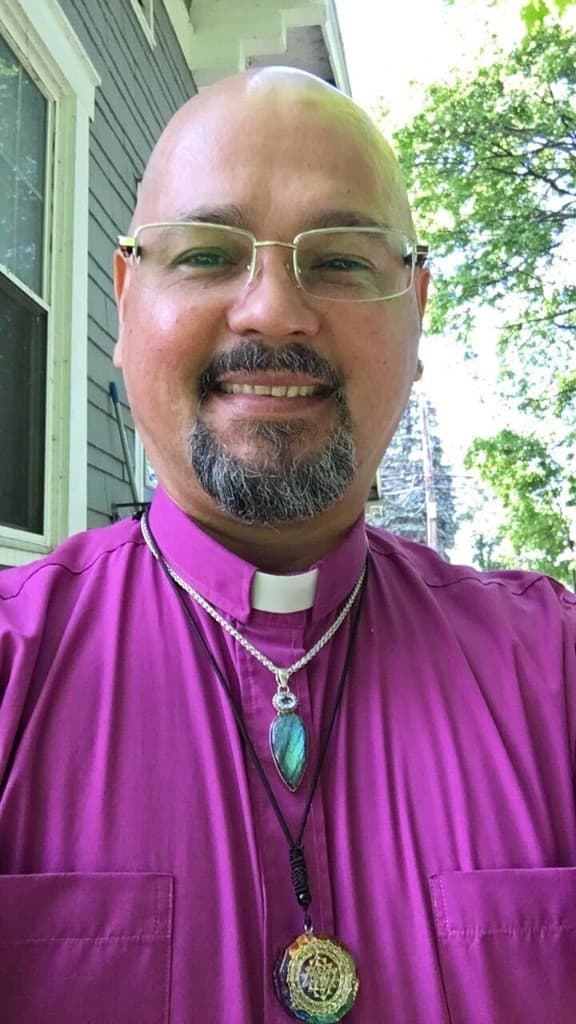 Christopher Byrne smiling outdoors, wearing a purple clerical collar and spiritual jewelry